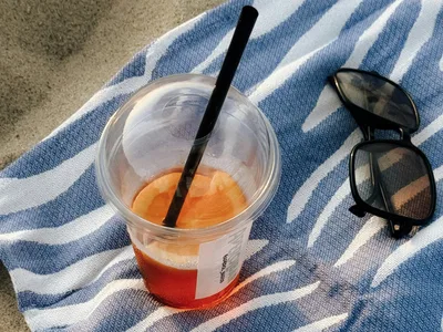 A spritz in a plastic cup and a pair of sunglasses on a striped blue-and-white towel in the sand.